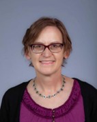 A woman with short brown hair and glasses, wearing a purple top and a beaded necklace, smiles at the camera against a plain background, representing the Plant Center at the upcoming Spring Symposium 2026.