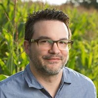 Man with short dark hair, glasses, and a beard wearing a blue shirt, standing outdoors in front of tall green plants at the Plant Center during the Spring Symposium 2026.