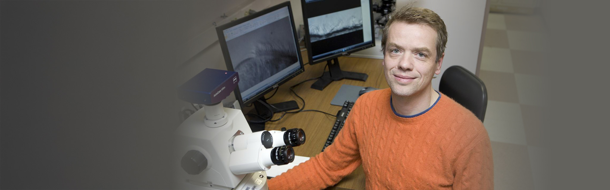 A person in an orange sweater sits at a desk with a microscope and two computer monitors displaying grayscale scientific images.