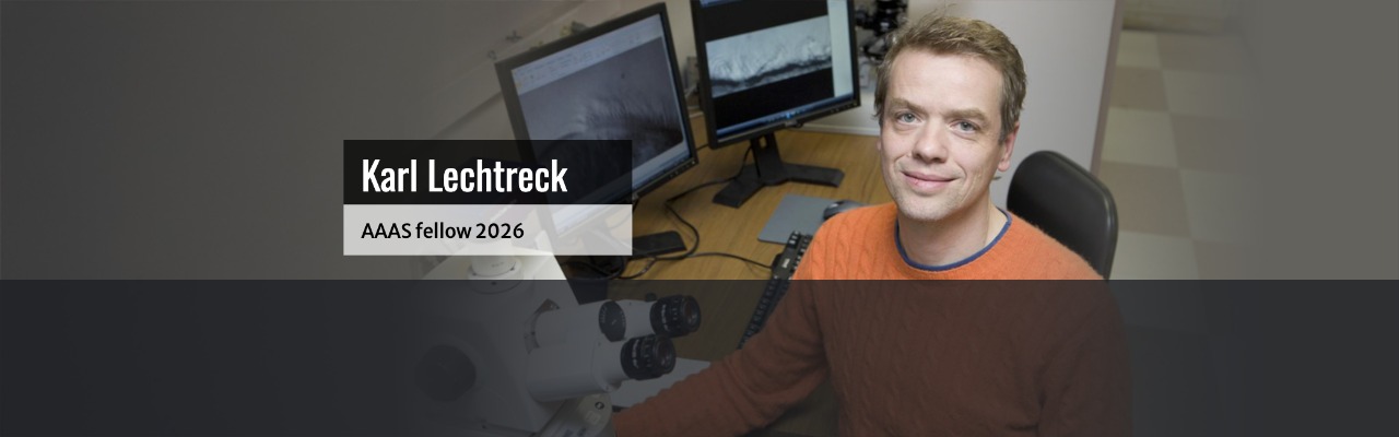 A man in an orange sweater sits at a desk with scientific equipment and computer monitors. Text reads: Karl Lechtreck, AAAS fellow 2026.