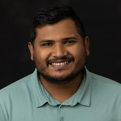 Man with short dark hair and beard, wearing a light green collared shirt, smiles at the camera against a plain dark background—this is Intiaz Amin Chowdhury.