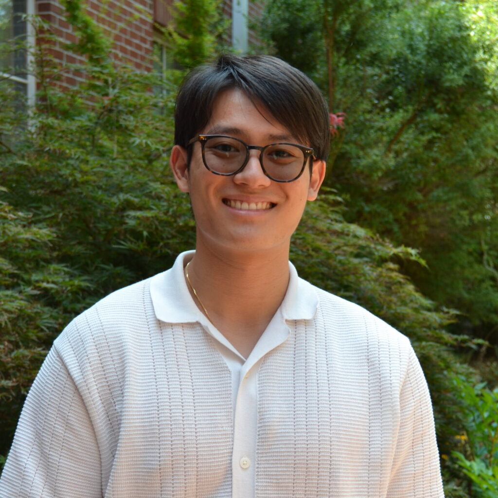 A person wearing glasses and a short-sleeve white shirt is standing outside in front of green plants and a brick wall, smiling at the camera.